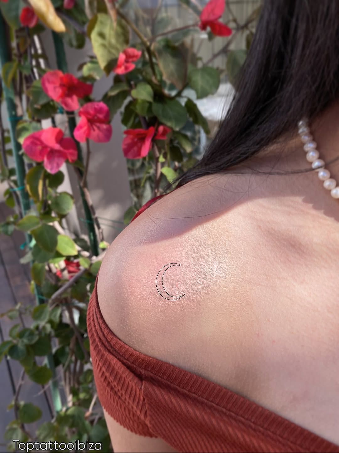 Fine-line crescent moon tattoo on a shoulder, bougainvillea in the background